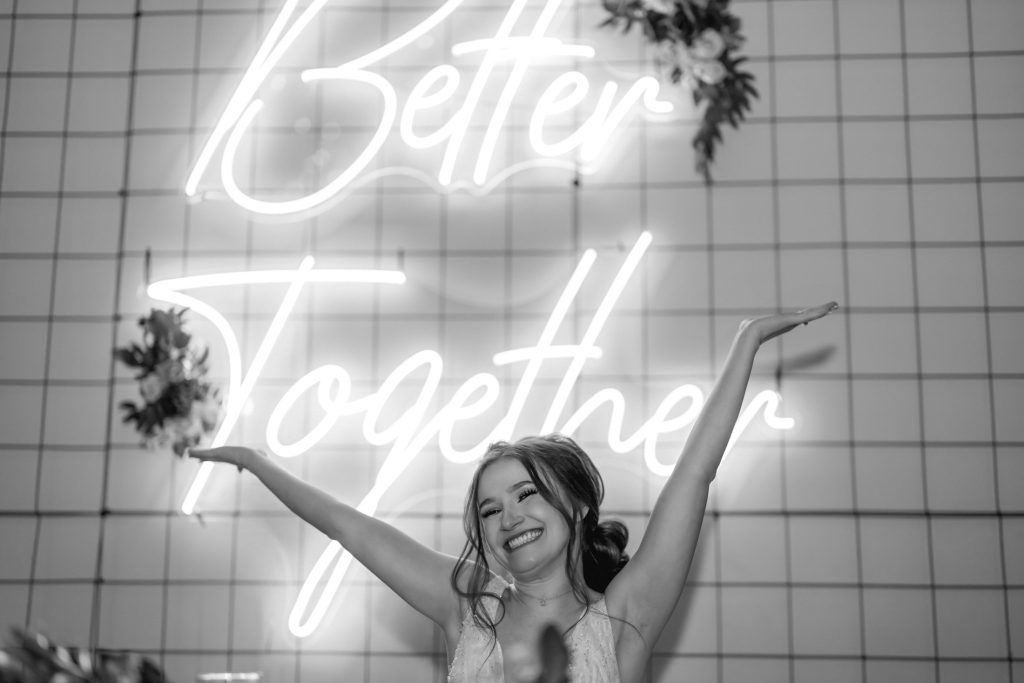 Bride celebrating with raised arms in front of neon Better Together sign black and white
