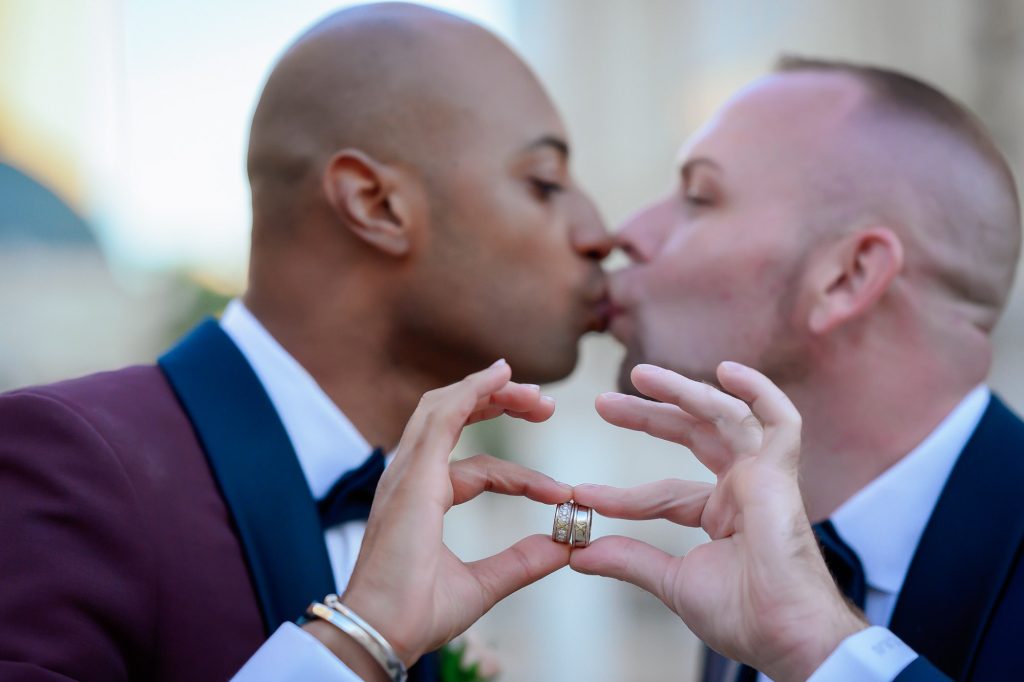 Same-sex wedding grooms kissing while holding wedding rings in heart shape