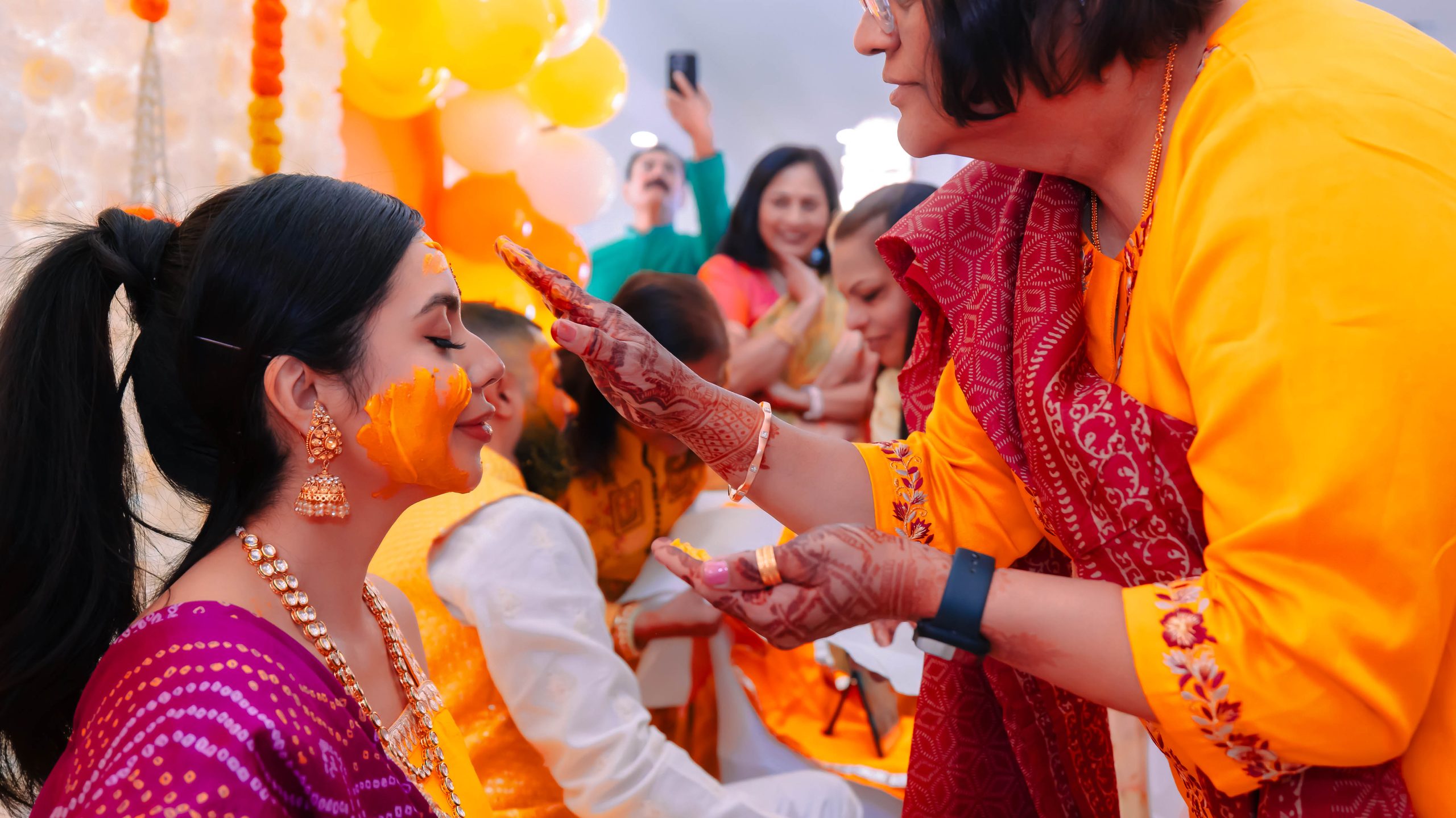 Bride receives a traditional haldi blessing during a vibrant Indian wedding ceremony surrounded by family and colorful decorations.