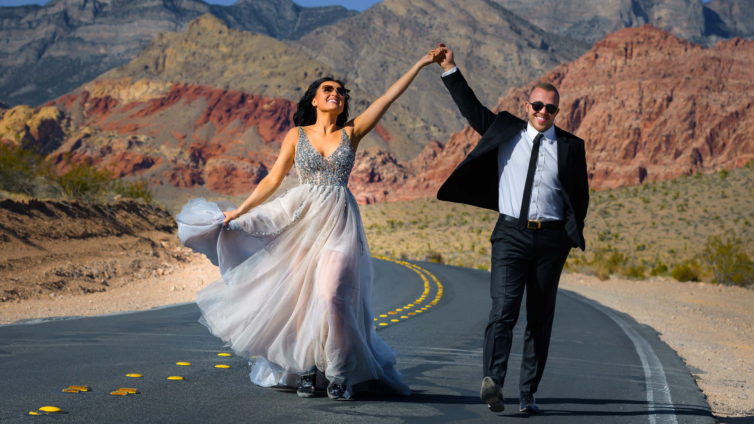 Newlywed couple walking hand in hand along a desert road in Red Rock Canyon near Las Vegas with colorful mountain scenery in the background.