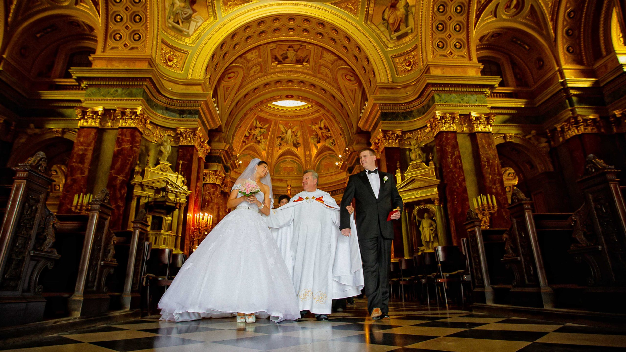 Wedding ceremony inside St. Stephen’s Basilica in Budapest featuring a bride and groom walking down the aisle beneath the cathedral’s ornate golden interior.