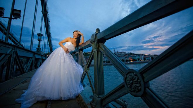 Bride posing in a wedding gown on Budapest’s Chain Bridge at dusk with the Danube River and city skyline in the background.