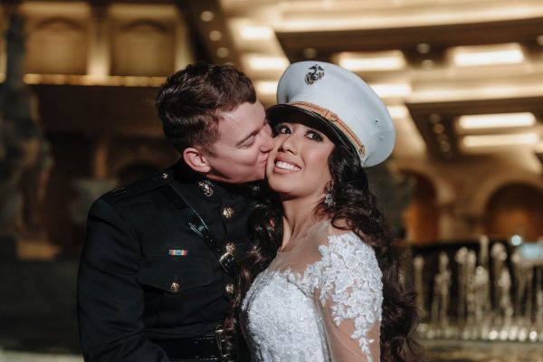 Bride wearing a U.S. Marine dress cap as her groom kisses her cheek during a romantic nighttime Caesars Palace wedding portrait in Las Vegas.