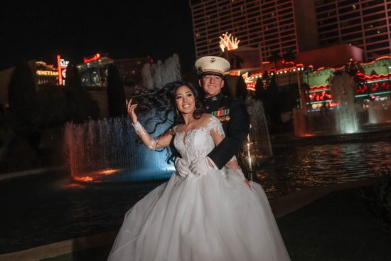 Luxury Caesars Palace wedding portrait at night with a U.S. Marine groom embracing his bride beside the illuminated fountains on the Las Vegas Strip.