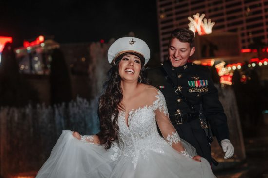 Luxury Caesars Palace wedding portrait at night with a smiling bride wearing a U.S. Marine dress cap as her groom looks at her beside the illuminated fountains.