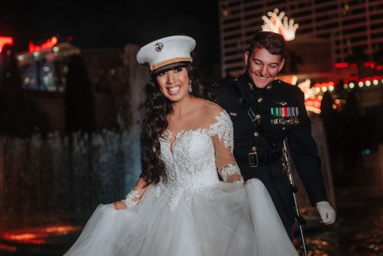 Luxury Caesars Palace wedding portrait at night featuring a smiling bride wearing a U.S. Marine dress cap with her groom beside the illuminated fountains in Las Vegas.