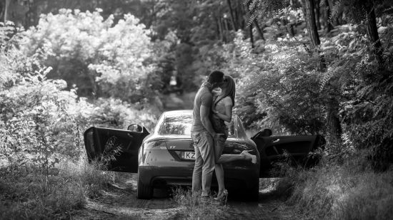 Black and white engagement photo of a couple kissing beside a car on a forest road in Hungary.